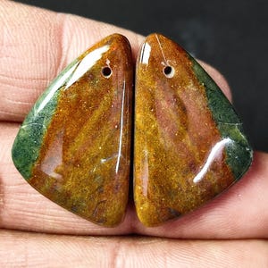 Puede incluir: Par de cabujones de piedra preciosa verde, marrón y naranja con agujeros perforados. Los cabujones son de forma triangular y tienen un acabado pulido.