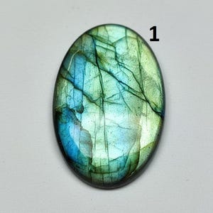 Può includere: Pietra preziosa di labradorite di forma ovale con superficie lucida. La pietra mostra un mix di colori blu, verde e turchese con venature interne. Il numero "1" è stampato in nero in alto a destra.
