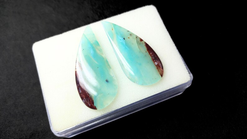 May include: Two teardrop-shaped cabochons of blue opal with brown inclusions. The stones are set in a white foam display box.