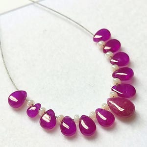 May include: A necklace featuring teardrop-shaped, magenta-colored beads interspersed with small, white beads. The beads are strung on a thin, silver-colored wire, creating a delicate and elegant design. The necklace is displayed on a white surface.