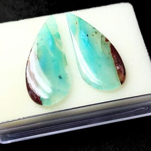 May include: Two teardrop-shaped cabochons of blue opal with brown and white inclusions. The stones are displayed in a white foam-lined box.