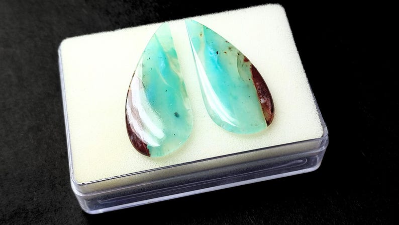 May include: Two teardrop-shaped cabochons of blue opal with brown and white inclusions. The stones are displayed in a white foam-lined box.