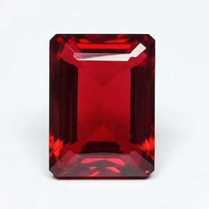 Puede incluir: Una gema roja oscura, tallada en forma rectangular. La gema tiene una superficie pulida y está sobre un fondo blanco. El corte de la gema crea múltiples facetas que reflejan la luz, realzando su brillo.