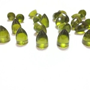 Puede incluir: Una colecci&oacute;n de cabujones de piedras preciosas verdes, cada uno con una forma puntiaguda en forma de l&aacute;grima. Las piedras est&aacute;n dispuestas sobre una superficie blanca.