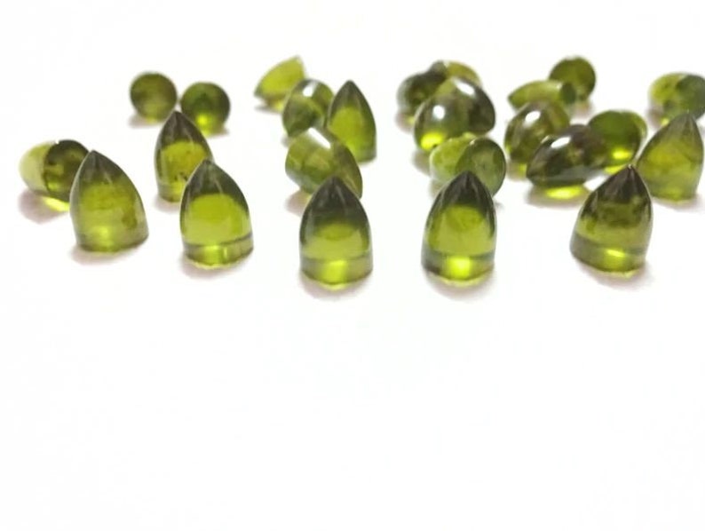 Puede incluir: Una colecci&oacute;n de cabujones de piedras preciosas verdes, cada uno con una forma puntiaguda en forma de l&aacute;grima. Las piedras est&aacute;n dispuestas sobre una superficie blanca.