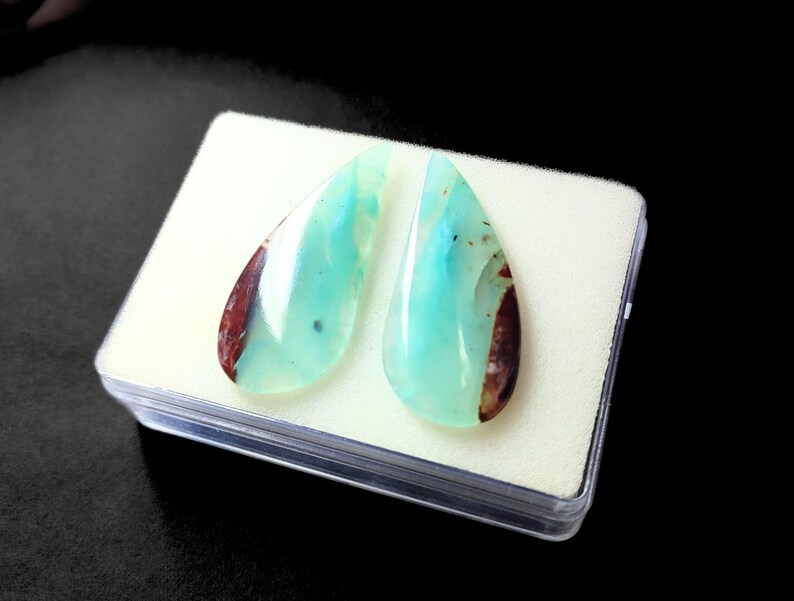 May include: Two teardrop-shaped cabochons of green opal with brown and red inclusions. The stones are displayed in a clear plastic box with white foam padding.