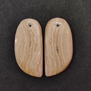 May include: Two polished, oval-shaped stone pendants. The pendants are a light beige colour with darker brown and tan veining patterns. Each pendant has a small hole drilled at the top for stringing.