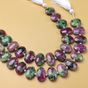 May include: Two strands of faceted gemstone beads in green and purple, with white tassels. The beads are irregularly shaped and polished. The background is a plain, light yellow colour.