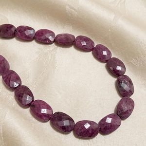 Puede incluir: Un collar con gemas facetadas de color rosa intenso, de forma ovalada. Las cuentas están ensartadas, creando un bucle continuo. Las gemas tienen una superficie pulida y reflectante, sobre una tela color crema.