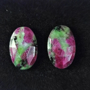 Könnte beinhalten: Zwei ovale Edelsteine mit Facettenschliff. Die Steine zeigen eine Mischung aus Farben, darunter tiefes Rosa, Grün und Schwarz. Die Edelsteine sind vor einem dunklen Hintergrund platziert.