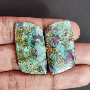 Peut inclure: Deux cabochons de pierres précieuses polis. Chaque pierre présente une matrice mouchetée de vert d'eau et de brun avec des inclusions rouge foncé. Les pierres sont rectangulaires avec un bord incurvé. Le fond est de couleur neutre.