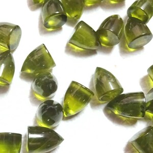 Puede incluir: Una colecci&oacute;n de 20 cabujones de piedras preciosas verdes, cada uno con una punta puntiaguda y una superficie lisa y pulida. Los cabujones est&aacute;n dispersos sobre un fondo blanco.