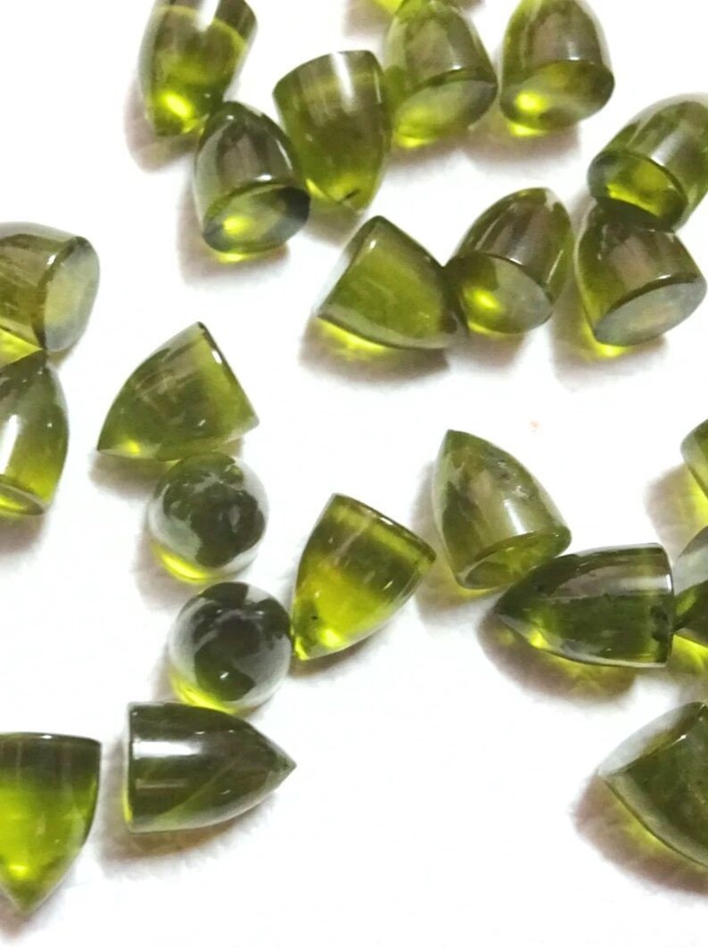 Puede incluir: Una colecci&oacute;n de 20 cabujones de piedras preciosas verdes, cada uno con una punta puntiaguda y una superficie lisa y pulida. Los cabujones est&aacute;n dispersos sobre un fondo blanco.