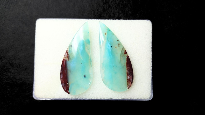 May include: Two teardrop-shaped cabochons of blue-green opal with brown inclusions. The stones are set in a white foam display.