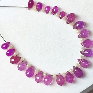 May include: A strand of faceted, teardrop-shaped, magenta-colored gemstones interspersed with small, white, round beads. The gemstones are strung on a thin, black thread, and are arranged in a curved line against a white background.