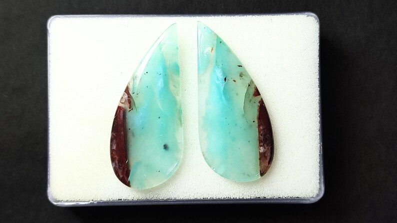 May include: A pair of teardrop-shaped cabochon gemstones with a blue-green color and brown inclusions. The stones are displayed in a white box.
