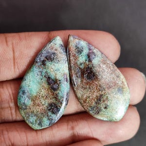 Peut inclure: Deux cabochons en forme de goutte présentant une surface marbrée avec un mélange d'inclusions bleu sarcelle vif, brun terreux et violet profond. Les pierres ont une finition polie et lisse qui souligne les variations de couleurs naturelles.