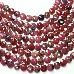 Puede incluir: Primer plano de varios hilos de cuentas de piedras preciosas facetadas, redondas y de color rojo oscuro. Las cuentas tienen un acabado brillante y pulido y están ensartadas. Cada hilo termina con una borla blanca. Las cuentas tienen aproximadamente 0,8 cm de diámetro.