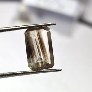 以下が含まれることがあります： 中央を垂直に走るダークブラウンの針状のインクルージョンを含む、透明で半透明のボディを持つ長方形のファセットカットされた宝石。石は金属製のピンセットで保持されており、磨かれた反射面が特徴です。