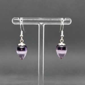 May include: A pair of dangle earrings featuring teardrop-shaped purple and white fluorite stones. The earrings have silver-colored hooks and caps. The stones are suspended from the hooks, creating a delicate and elegant look.