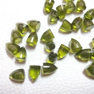 Puede incluir: Una colecci&oacute;n de cabujones de piedras preciosas verdes en forma de gota puntiaguda. Las piedras est&aacute;n esparcidas sobre una superficie blanca.