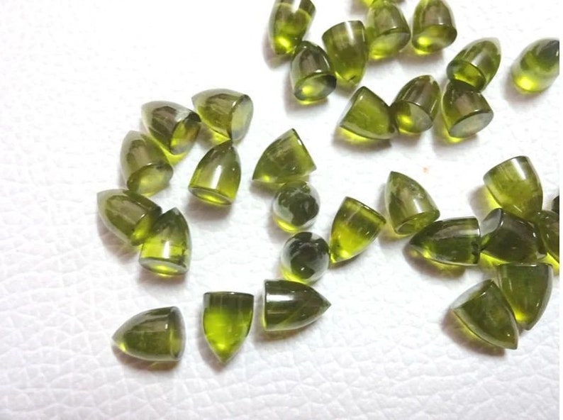 Puede incluir: Una colecci&oacute;n de cabujones de piedras preciosas verdes en forma de gota puntiaguda. Las piedras est&aacute;n esparcidas sobre una superficie blanca.