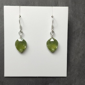 以下が含まれることがあります： 洋梨型の、ファセット加工が施されたオリーブグリーンの宝石をあしらったイヤリングのペア。宝石は、銀色の湾曲したイヤーフックから吊り下げられています。イヤリングは白い背景に表示されています。