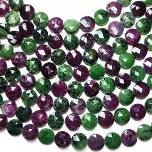 Puede incluir: Primer plano de cuentas de piedras preciosas facetadas en tonos morado oscuro y verde. Las cuentas redondas están ensartadas, con una borla amarilla brillante en el lado izquierdo. Las piedras preciosas tienen un aspecto brillante y pulido.