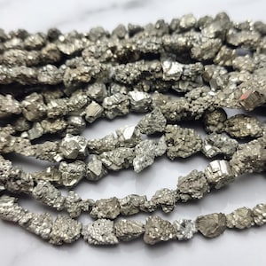 May include: A strand of raw pyrite beads featuring metallic, brassy-gold, and silver-toned mineral nuggets with natural, irregular cubic crystalline structures. The beads are strung together, showcasing a reflective, textured, and multifaceted surface.