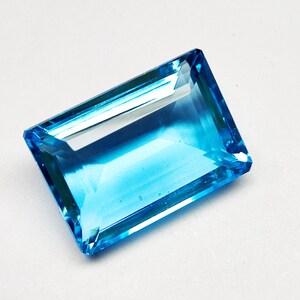 Pode incluir: Uma gema retangular e facetada com um tom azul claro vibrante. As facetas da gema refletem a luz, criando um efeito cintilante. A gema está sobre um fundo branco, realçando sua cor e clareza.
