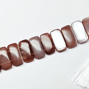May include: A strand of rectangular, smooth, polished beads in shades of mauve, brown, and iridescent white, arranged in a linear sequence and finished with a single, long, white decorative tassel attached by a small silver-toned metal cap.