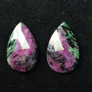 Könnte beinhalten: Zwei tropfenförmige Edelsteine mit facettierten Oberflächen. Die Steine weisen eine lebendige Mischung aus Farben auf, darunter tiefes Magenta, Schwarz und Grün. Die Edelsteine sind vor einem dunklen Hintergrund platziert, der ihre einzigartigen Farbmuster und den Schliff hervorhebt.