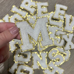 May include: A close-up of a white felt letter M with gold glitter trim. The letter is surrounded by other letters, all with gold glitter trim.