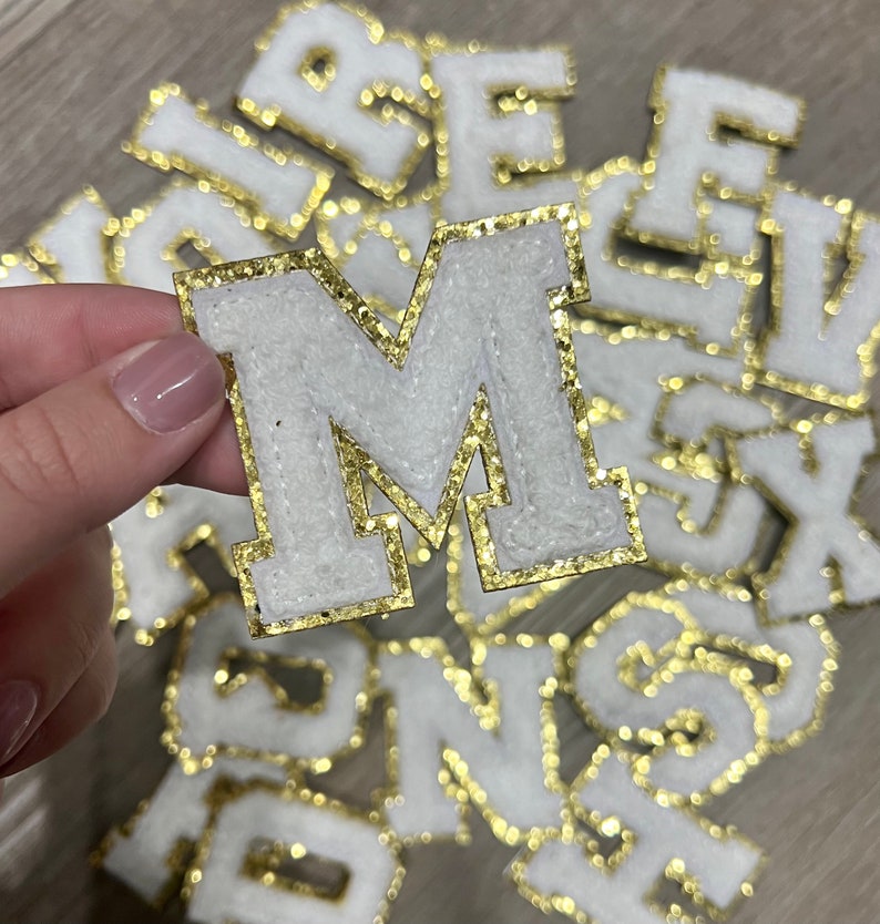 May include: A close-up of a white felt letter M with gold glitter trim. The letter is surrounded by other letters, all with gold glitter trim.