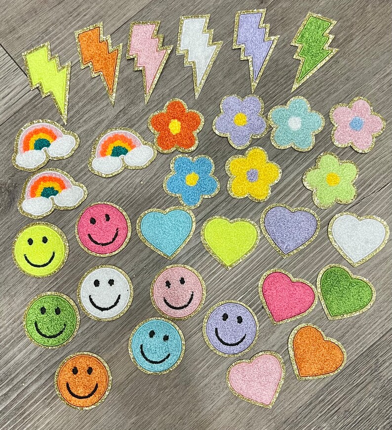May include: A collection of colorful iron-on patches featuring various designs, including rainbows, smiley faces, hearts, flowers, and lightning bolts. The patches have a gold glitter outline and are perfect for adding a touch of personality to clothing, bags, or other accessories.