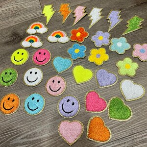 May include: Assortment of colorful iron-on patches featuring various designs, including smiley faces, hearts, lightning bolts, rainbows, and flowers. The patches are all made of glitter fabric and have a gold outline.