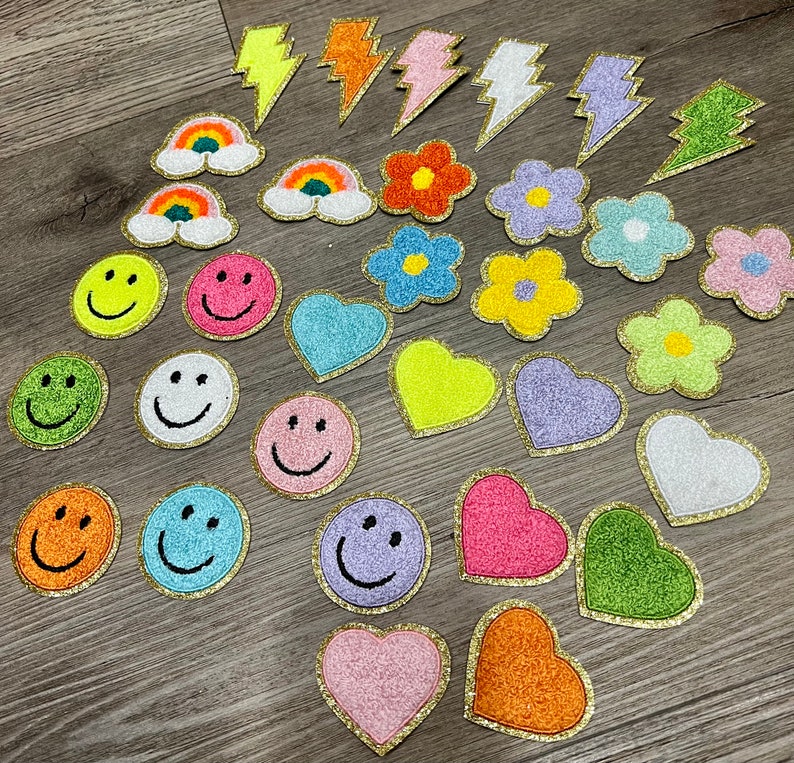 May include: Assortment of colorful iron-on patches featuring various designs, including smiley faces, hearts, lightning bolts, rainbows, and flowers. The patches are all made of glitter fabric and have a gold outline.