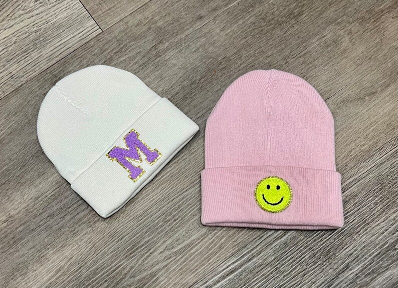 May include: Two knit beanies, one white with a purple "M" and one pink with a yellow smiley face.