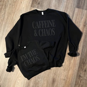 May include: Two black sweatshirts with the phrases "Caffeine & Chaos" and "I'M THE CHAOS" printed on them. The sweatshirts are displayed on a wooden surface. The larger sweatshirt is an adult size, and the smaller one is a child's size.
