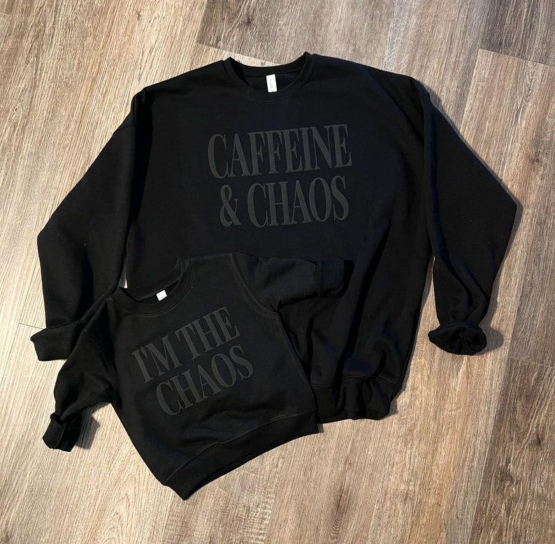May include: Two black sweatshirts with the phrases "Caffeine & Chaos" and "I'M THE CHAOS" printed on them. The sweatshirts are displayed on a wooden surface. The larger sweatshirt is an adult size, and the smaller one is a child's size.