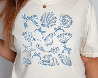 Op de afbeelding: Een wit T-shirt met een blauw schelpontwerp. Het ontwerp bevat verschillende schelpen, zeesterren en strikken.