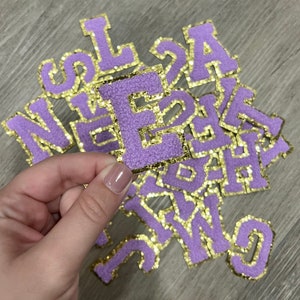 May include: A pile of purple felt letters with gold glitter trim. The letters are in various sizes and are arranged in a random order. The letter "E" is being held up by a hand.