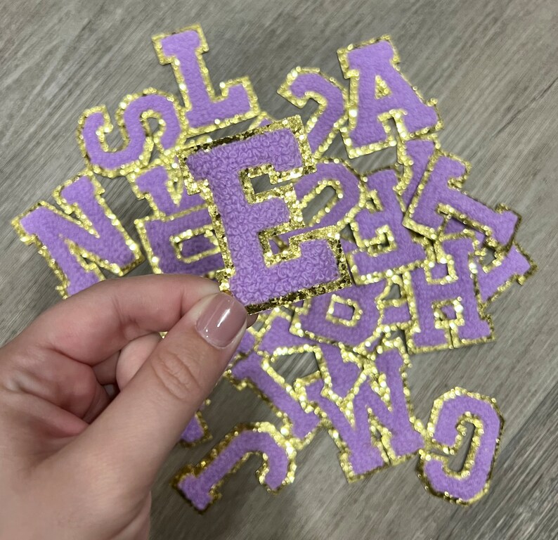 May include: A pile of purple felt letters with gold glitter trim. The letters are in various sizes and are arranged in a random order. The letter "E" is being held up by a hand.