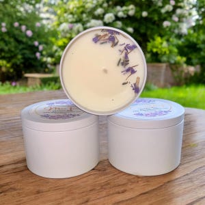 Puede incluir: Tres tarros de velas blancas con flores de lavanda incrustadas en la cera. Los tarros tienen una etiqueta blanca con el texto "Morning Glory" y una dirección de sitio web.