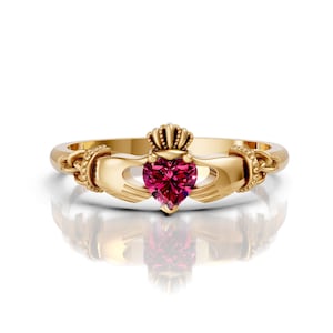 Puede incluir: Un anillo Claddagh celta de oro con una piedra preciosa roja en forma de corazón. El anillo presenta dos manos entrelazadas, simbolizando la amistad y el amor, con una corona sobre las manos, que representa la lealtad.