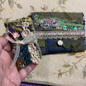 Könnte beinhalten: Ein kleines rechteckiges Stofftäschchen und ein passendes gefaltetes Stoffheft, beide in Patchwork-Optik mit olivgrünen, marineblauen und violetten Tönen, verziert mit Spitzenbesatz, irisierenden grünen Akzenten und kleinen floralen Details.