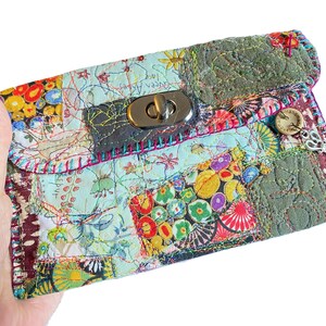 Könnte beinhalten: Eine Patchwork-Clutch mit silbernem Verschluss. Die handgefertigte Tasche hat eine Vielzahl von bunten Stoffquadraten mit floralen und abstrakten Mustern. Der Überschlag wird mit einem silbernen Verschluss gesichert. Ein kleiner Holzknopf und ein Anhänger sind an der Seite befestigt.