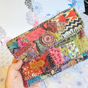 Peut inclure: Pochette patchwork faite à la main, aux couleurs vives : rouge, rose, vert et violet. Fermeture par un gros bouton. Une petite étiquette en cuir porte l'inscription "Handmade". Motifs floraux et géométriques se côtoient.