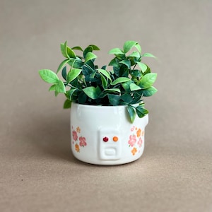 May include: A small, off-white ceramic planter shaped like a rice cooker, adorned with orange and pink floral designs. The planter is filled with vibrant green artificial foliage. The design includes a rectangular panel with two orange dots and a small rectangle.