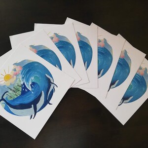 May include: A set of eight blank greeting cards featuring a watercolor illustration of a whale breaching in a wave with a sun and trees in the background. The cards are white with a blue and green watercolor design.